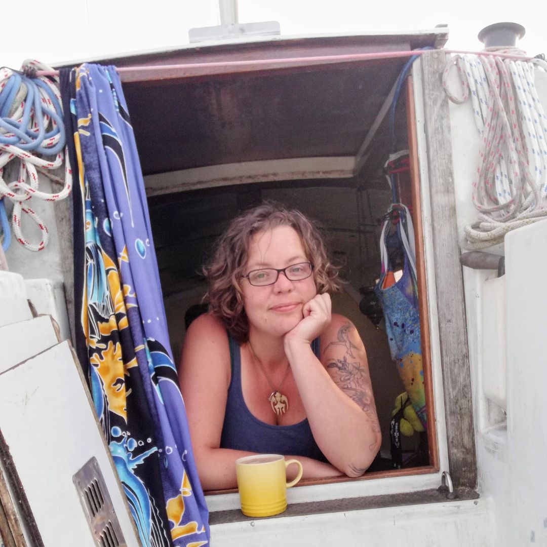 Mad Jessie looking out window of a boat house with a colorful patterned fabric hanging to hr side